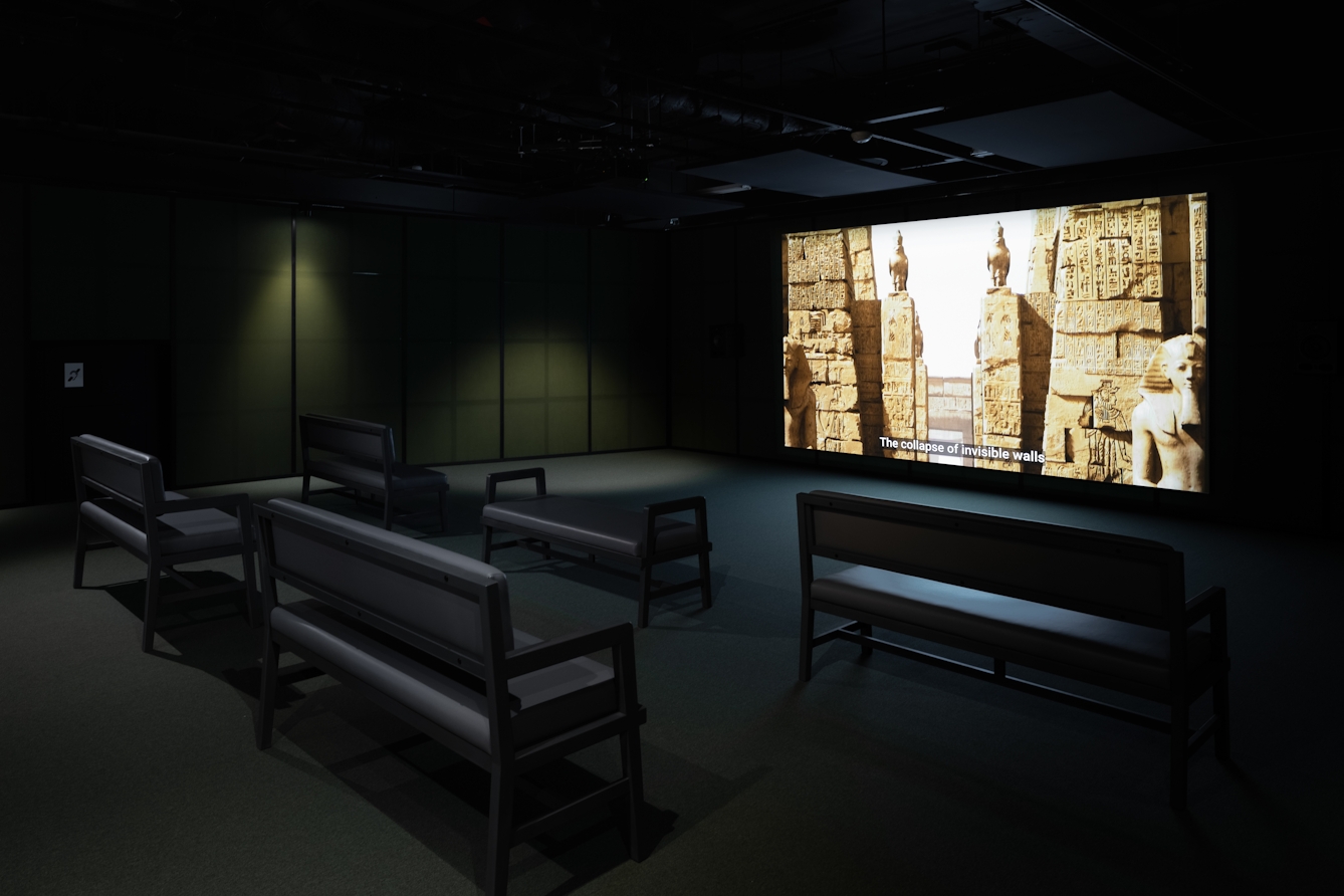 Photograph of a dark exhibition space featuring a projected video with subtitles on a large screen. 5 bench seats are arranged facing the screen. The walls of the room are a criss-cross of black wooden battens on grey fabric. The video shows a computer generated scene of an ancient Egyptian settlement, with towers, hieroglyphics and sphinxes. The subtitles below read 'The collapse of invisible walls'.