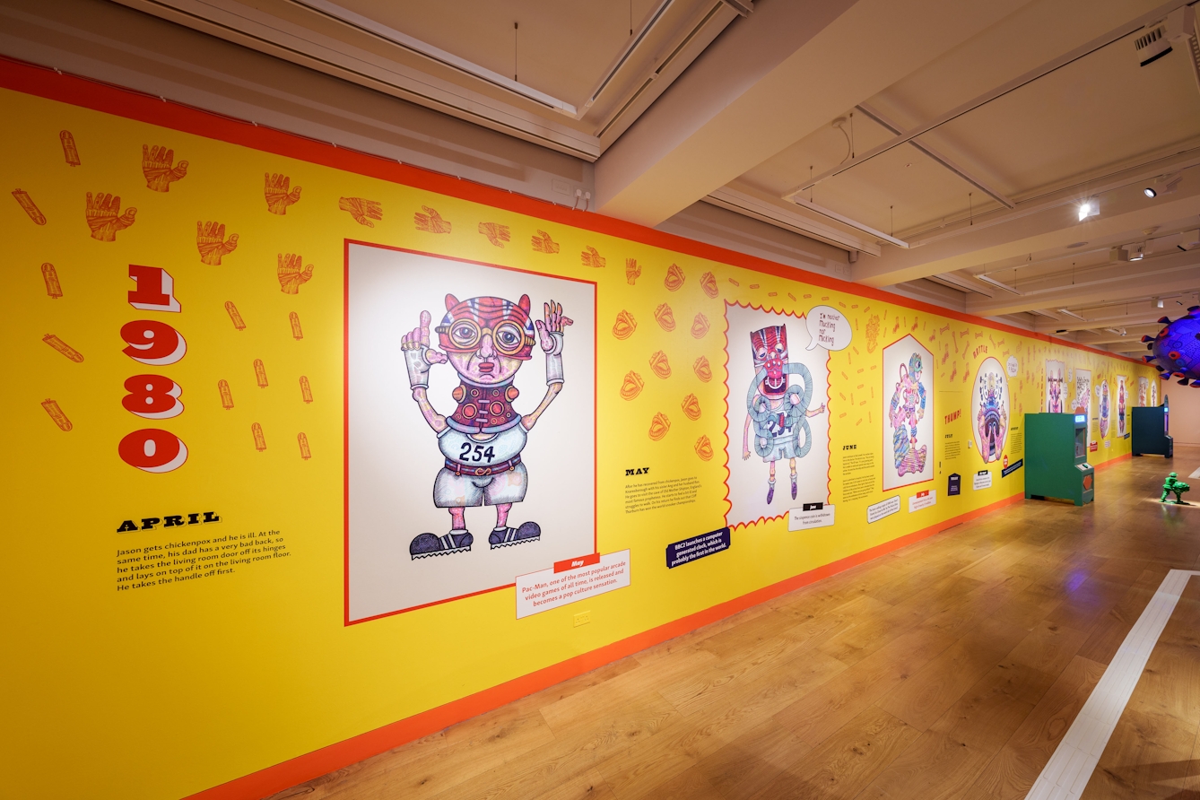 A long wall covered in brightly coloured wallpaper. The wallpaper shows drawings of fantastic figures and writing about the year 1980.