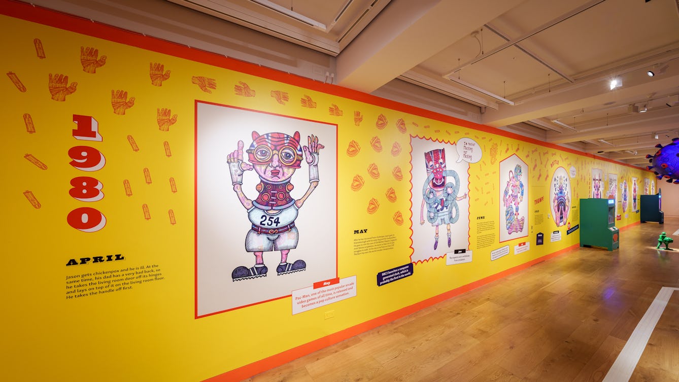 A long wall covered in brightly coloured wallpaper. The wallpaper shows drawings of fantastic figures and writing about the year 1980.