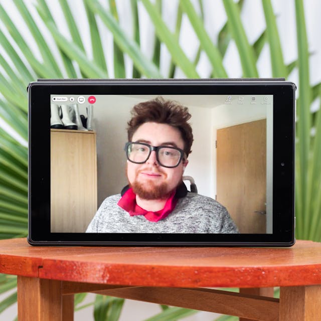 Photograph of a tablet standing on a wooden stool. On the tablet screen is a photographic portrait of Jamie Hale, wearing glasses, a grey sweater with a pink collar showing. Also, on the screen are video call icons "People", "Chat" and a red telephone icon. There are green decorative palm leaves displayed in the background behind the tablet which is next to a colourful folded blanket and the edge of a blue sofa with a yellow cushion.
