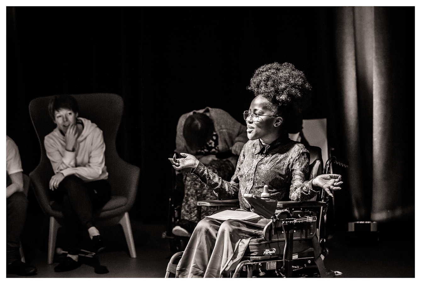 Black and white photograph with a warm tone. The image shows a young woman seated in a wheelchair against a black curtain and lit by a spotlight. She is performing to an audience which can be seen seated to her right int he background. She has been captured 'in action'.