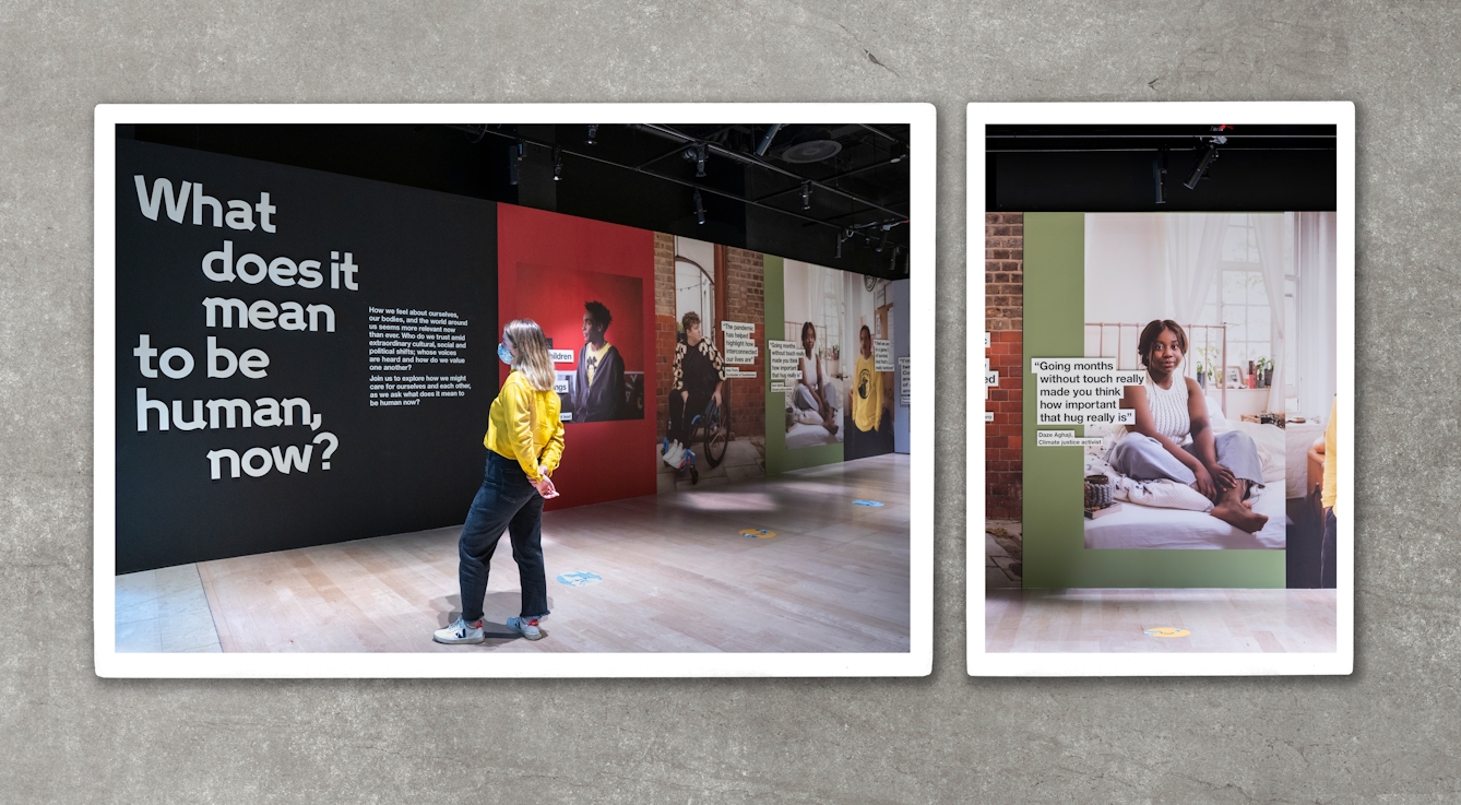 Photograph of two photographic colour prints resting on a grey, concrete textured surface, from directly above. The prints are slightly raise off the surface, casting a small shadow. The prints show a gallery installation. The image on the left shows an individual in a yellow top and dark trousers, wearing a face covering, who is reading an information panel on the gallery wall. In front of them are large title graphics with the words, 'What does it mean to be human, now?'. The gallery wall recedes into the background, where large portraits and more text can be seen. The image on the right shows one of the exhibition portraits of a woman sat on a bed looking to camera. Mounted on top of the print is the quote, 'Going months without touch really made you think how important that hug really is".