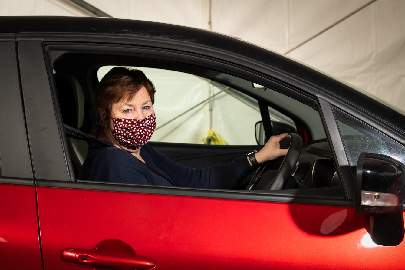 Photographic flash lit portrait through the open side window of a stationary car. The car is red and black and the front of the car is facing to the right. Sat in the driver's seat with her hand on the steering wheel is a woman, looking to the camera. She is wearing a floral maroon face covering. In the background is the out of focus inside of a white marquee.