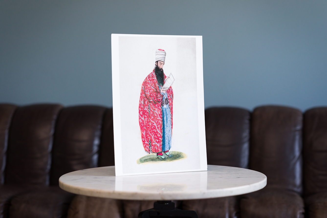 Photograph showing circular marble tabletop in front of a large brown sofa. Propped up on the tabletop is a print of an illustration of a Persian astrologer or mathematician from the Qajar period. The individual has a long black beard.