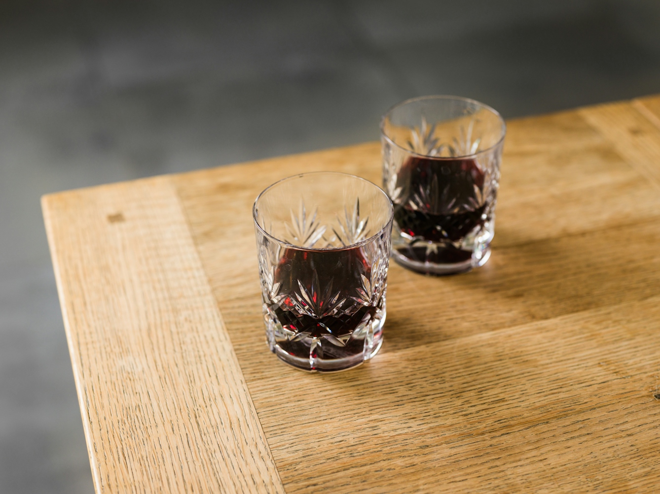 A photograph of a corner of a wooden table with two small tumbler-style glasses. The glasses contain red wine, and are filled below halfway. The glasses have detailed ridges on the outside in palmleaf-like patterns.