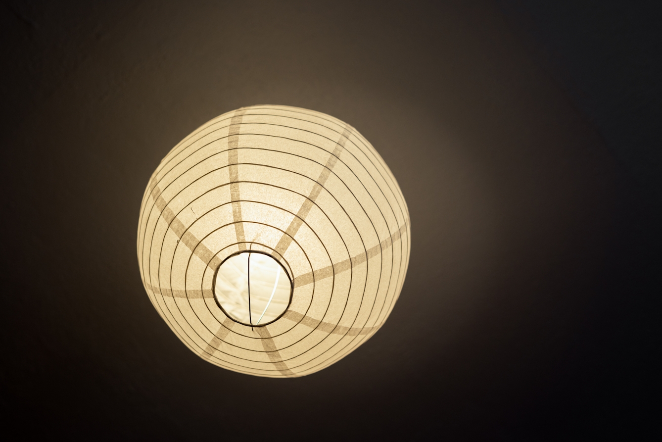 Photograph looking up at a spherical paper ceiling lampshade. The lamp is on, illuminating the lampshade and a patch of the ceiling. The lampshade is made of paper and a spiralling metal wire. The image has a warm tone to it. 