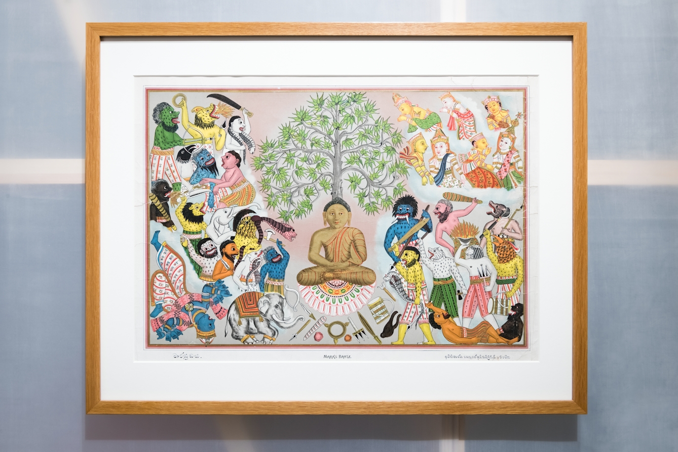 Photograph in an exhibition gallery showing a section of a translucent wall revealing the wooden structure behind, on which a framed print in a wooden frame has been hung. In a window mount within the frame is a colourful drawing of a Buddha, resisting the demons of Mara, who are attempting to prevent him from attaining enlightenment, as angels watch from above.