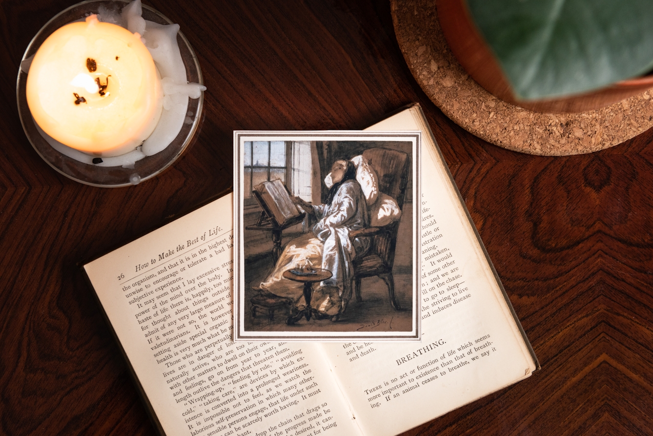 Photograph showing a candle, book and painting on a wooden table. 

The book is open and the chapter heading reads 'How to Make the Best of Life'. The painting shows a young woman sat in a chair with pillows, who is convalescing, reading a book by a window. 