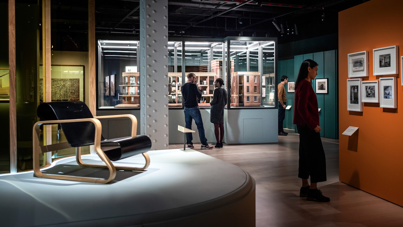Photograph of visitors exploring the exhibition, Living with Buildings at Wellcome Collection. Photograph shows an architectural model in a display case, an armchair and framed works hanging on the wall. 