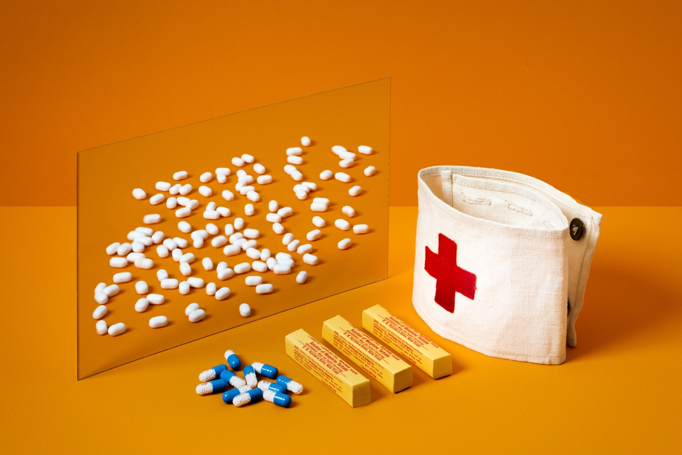 A photograph of a mirror on an orange background. In the foreground is a medic’s armband with a Red Cross, packages of painkillers and some loose pills. In the reflection of the mirror are dozens of white sugar pills.