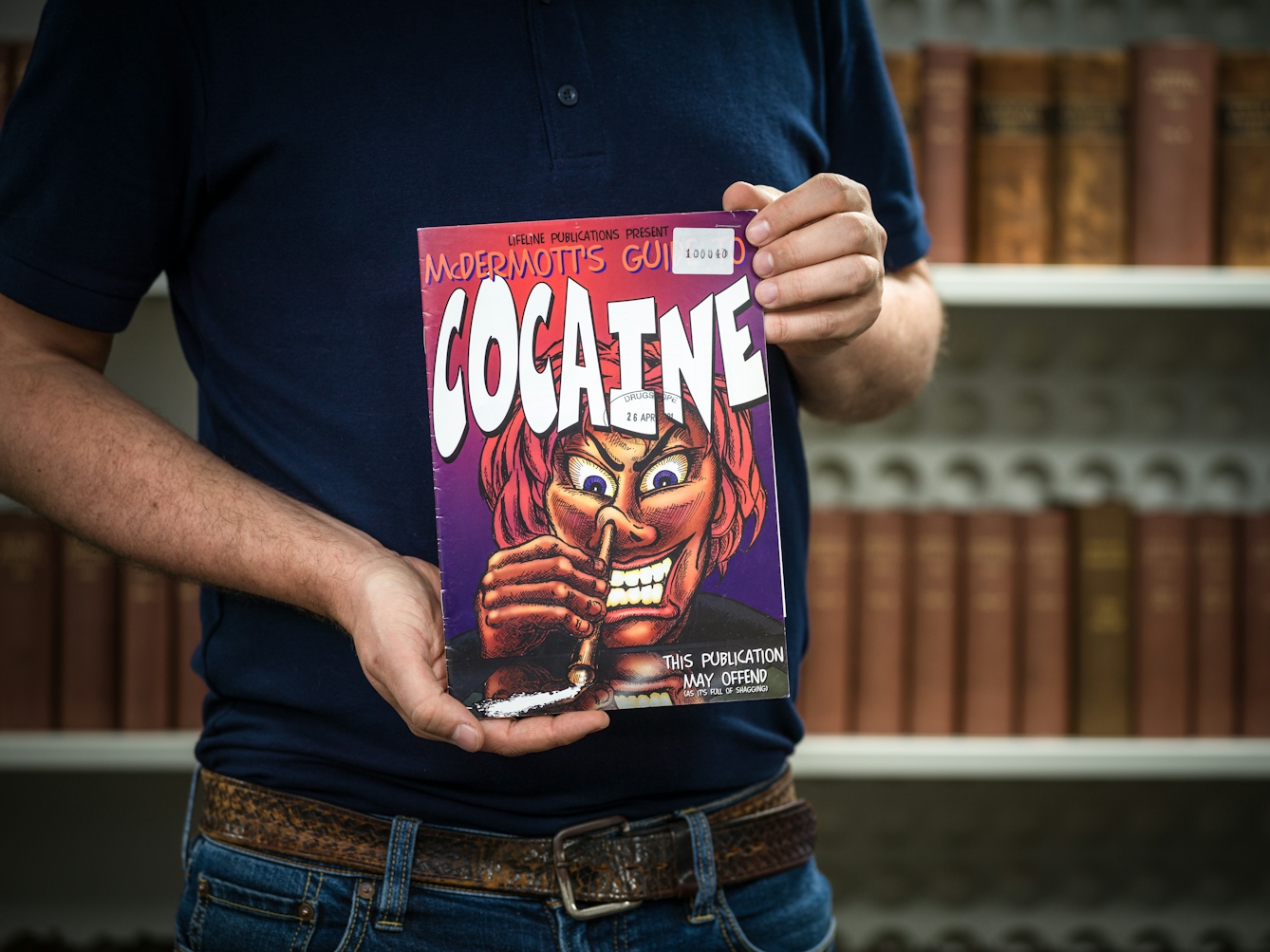 Photograph of the midriff of a man who is holding a pamphlet titled 'Cocaine, this publication may offend'. In the background are a set of out of focus shelves containing books.