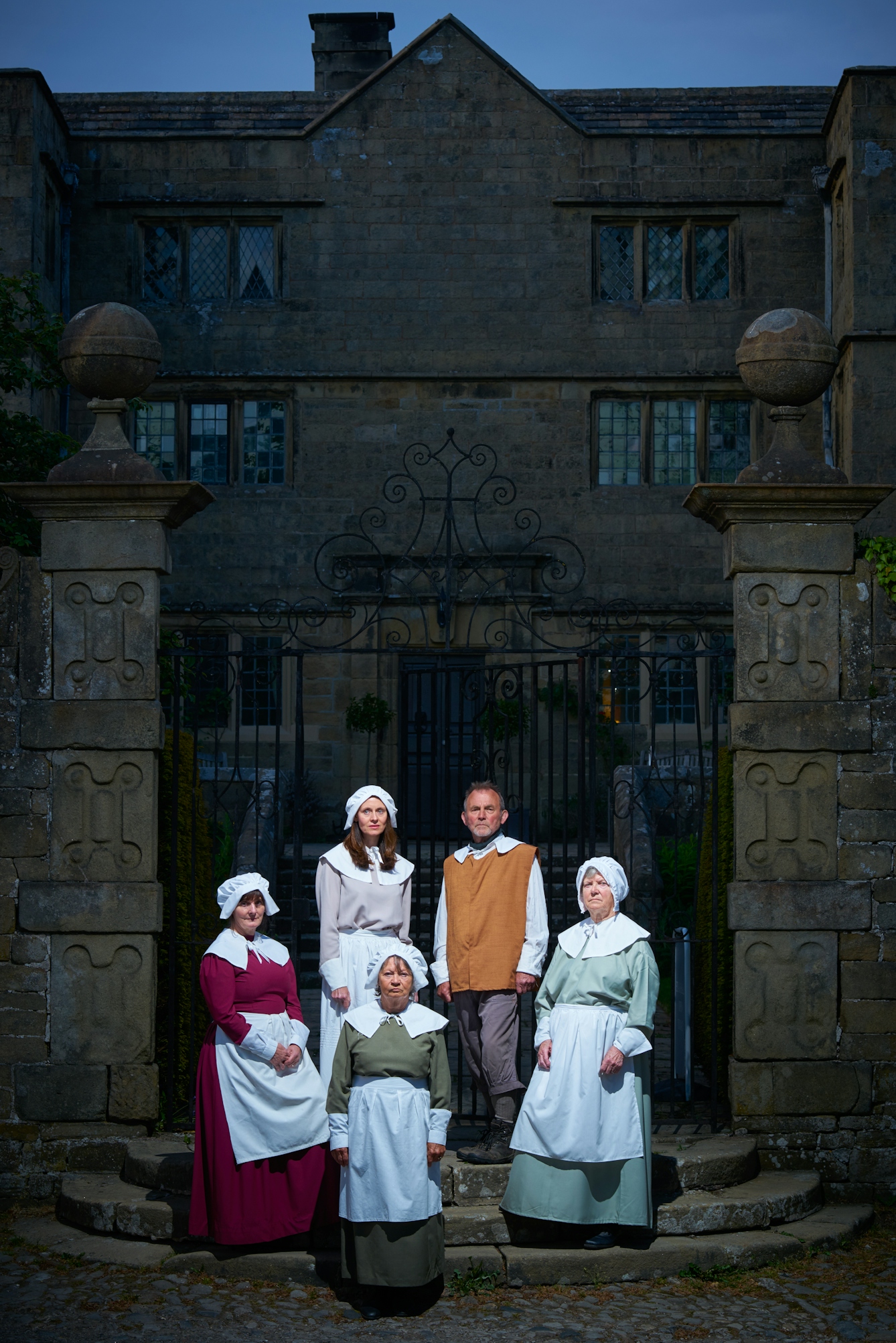 Photograph shot at dusk of the entrance to a stately home or hall. In a pool of light in the foreground is a group of 4 women and 1 man dressed in traditional 17 century clothes, looking towards the camera.