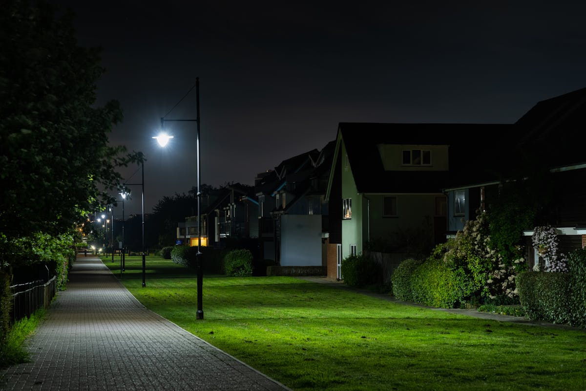 A photograph of a pathway at night which is lined by a row of street lights. The lights illuminate the trees and lawns next to the path as well as a row of houses, the gardens of which also has light cast on them.