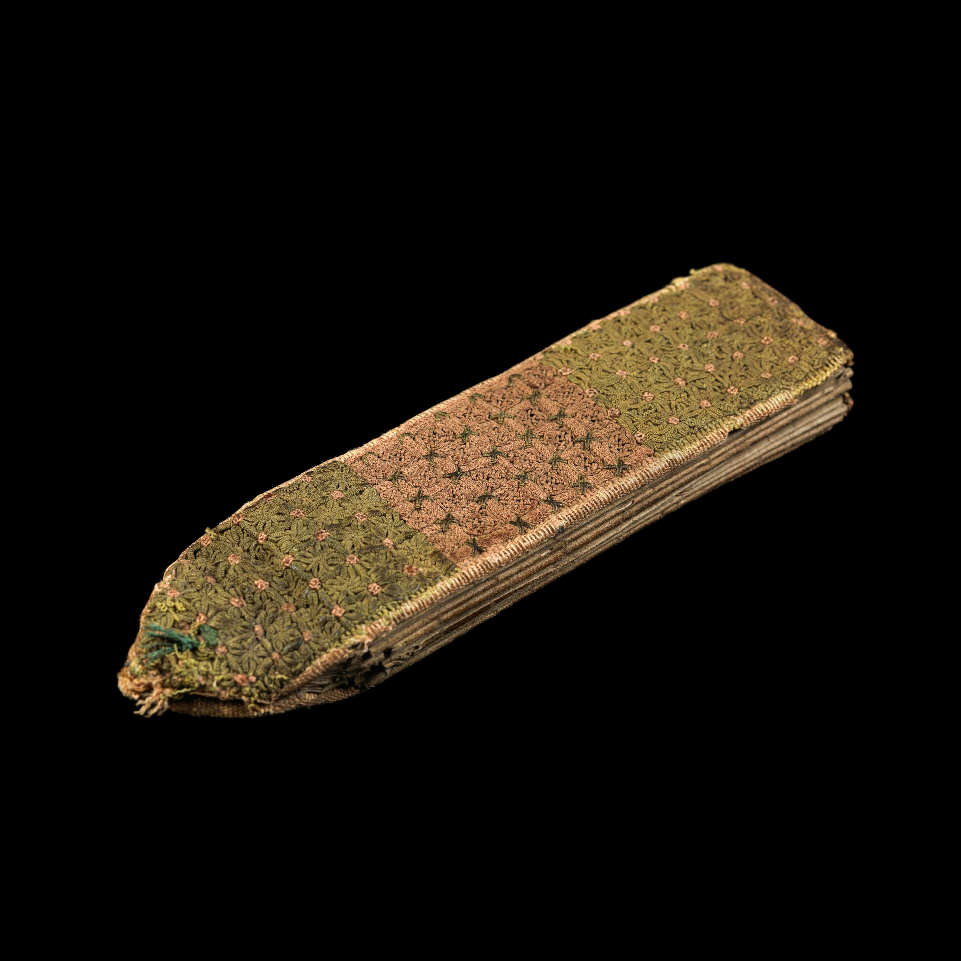 Photograph of a fragile folding almanac from the 15th century. The almanac is spotlit on a black background. It has an intricate woven green and pink fabric cover and folded internal pages. It is rectangular in shape, but with a triangular taper on the left short end.