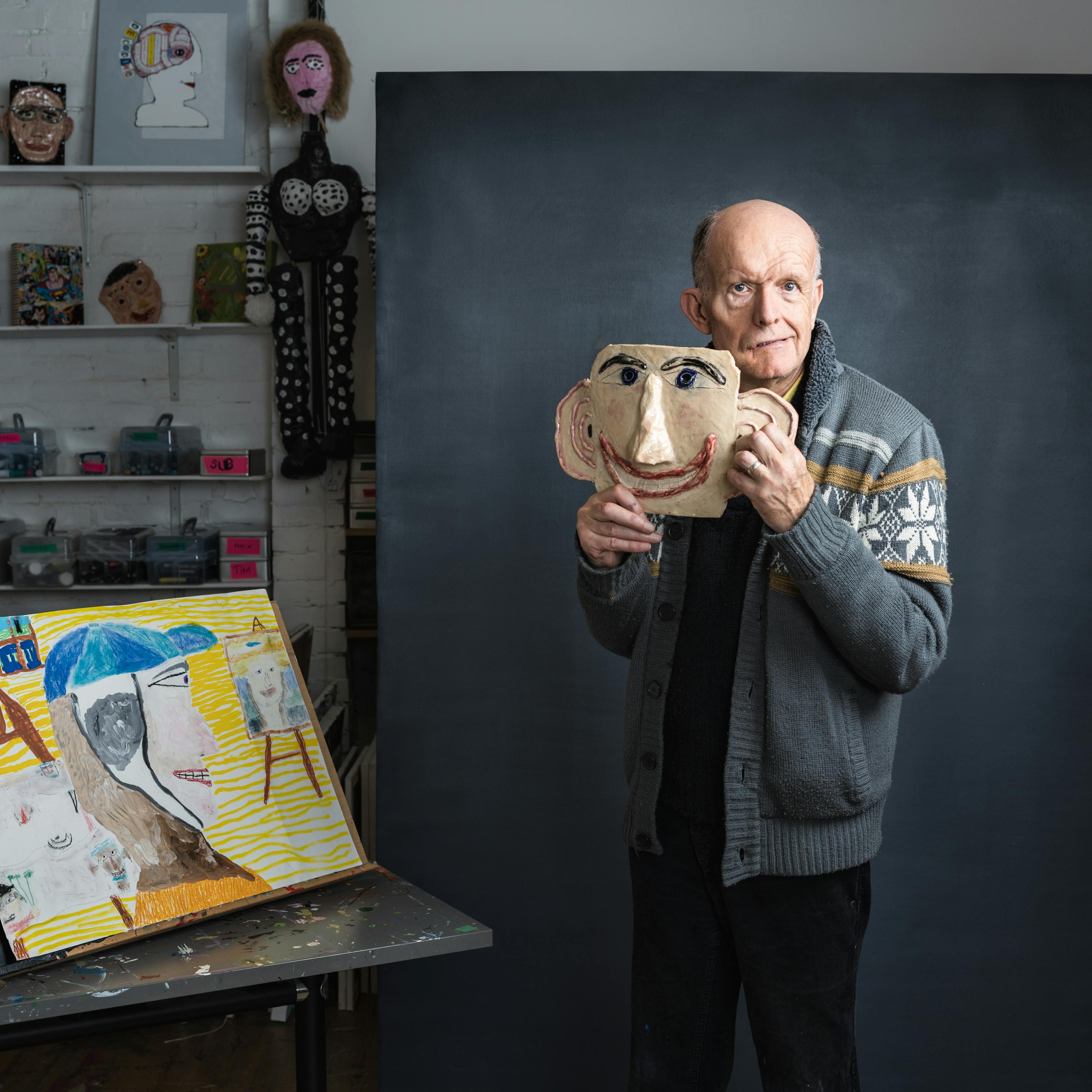 Photograph of stroke survivor and artist Chris Miller in an artist studio. A grey textured background frames the artist as he holds a ceramic of a head, which acts as a cartoon likeness of his own face. Miller’s gaze is directed towards the viewer. In the background of the studio the room is layered with artworks. To the left of the frame is a yellow painting on a table top easel.