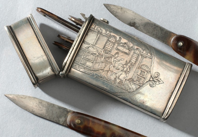 Photo of an engraved metal container with several blades inside and two blades either side of it.