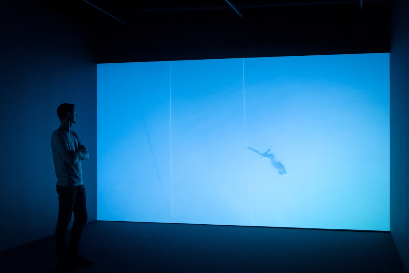 Photograph of a visitor exploring a gallery installation. He is standing in front of a large projection of a figure floating underwater. The room has a blue cast.