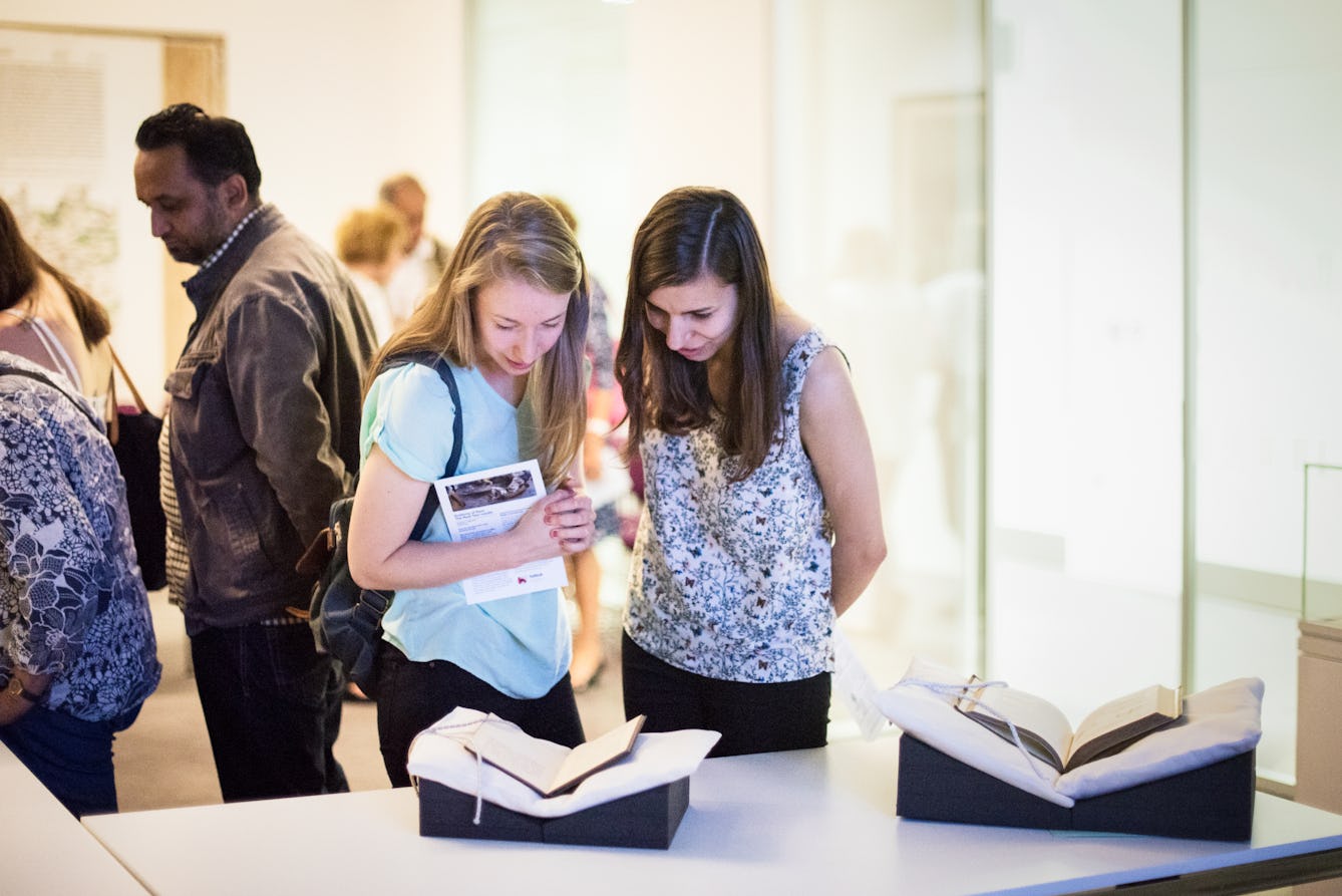 Photograph of two young women standing next to a table in a museum viewing room. On the tabletop in front of them are two open books, supported in book cradles. They are both looking down at one of the books, in discussion. Behind them are other visitors looking at other items.