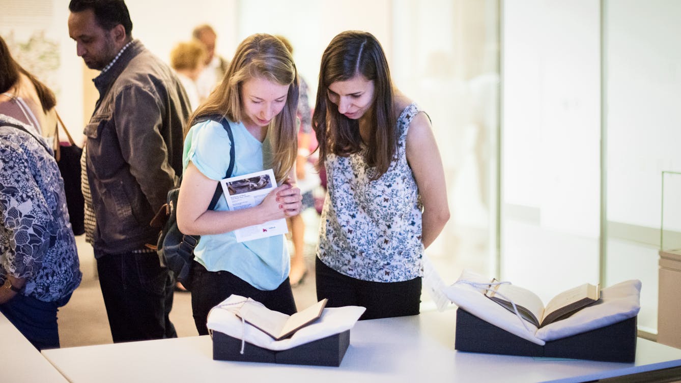 Photograph of two young women standing next to a table in a museum viewing room. On the tabletop in front of them are two open books, supported in book cradles. They are both looking down at one of the books, in discussion. Behind them are other visitors looking at other items.