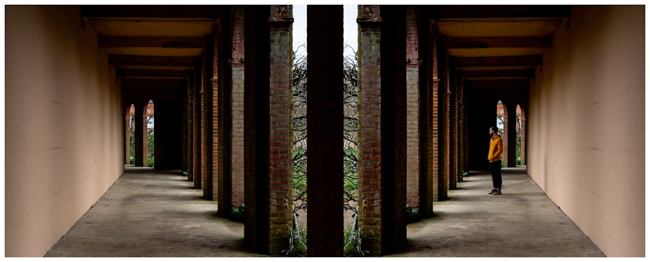Photographic panorama showing an arched brick walkway. The panorama is mirrored down its vertical centre line except that a small human figure in a yellow top standing in the archways appears in the right half, but not in the left half.
