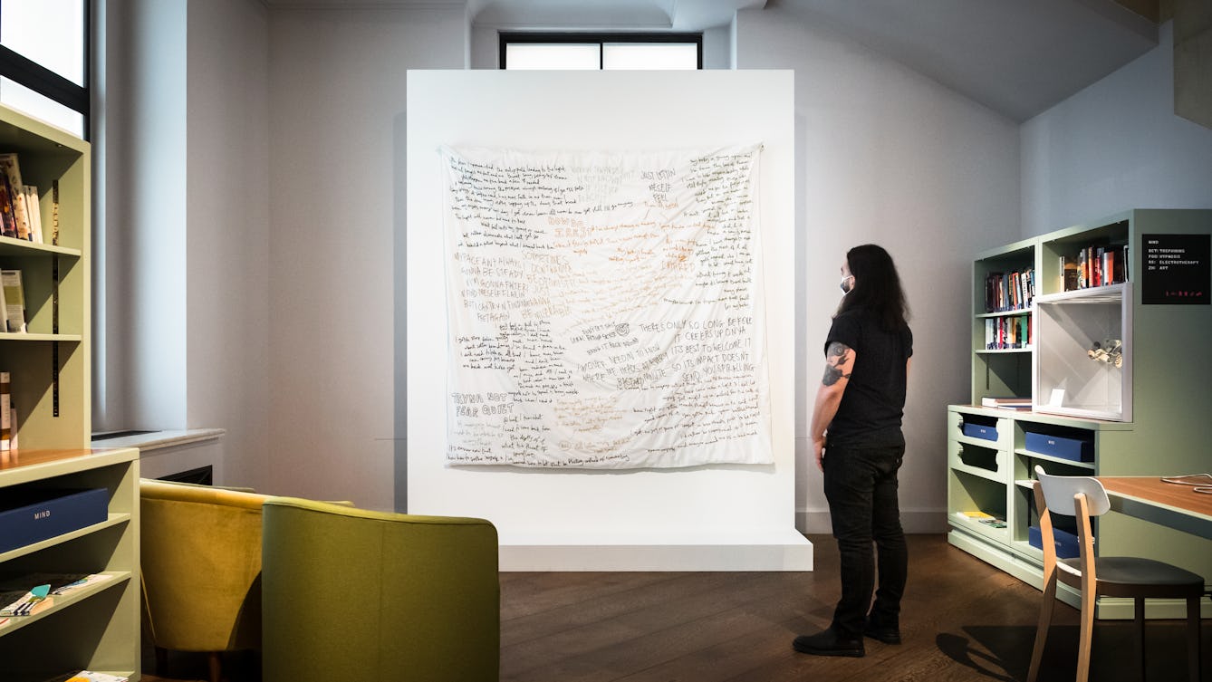 Photograph of a gallery installation showing a large white freestanding wall on which an artwork is. The artwork is a white sheet covered in handwritten text. Some of the text is black and some orange and red. A man wearing a face covering and black jeans and a t-shirt is standing to the right of the artwork looking at it. The surrounding gallery space contains, bookshelves, chairs and a table and a large window.
