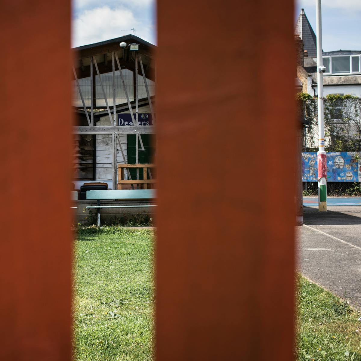 Photograph of an adventure playground, taken through a vertical slatted wooden fence. The vertical fence panels obscure much of the view, but through the gap are glimpses of a grass area, a concrete play area and a large pitched roof community building.
