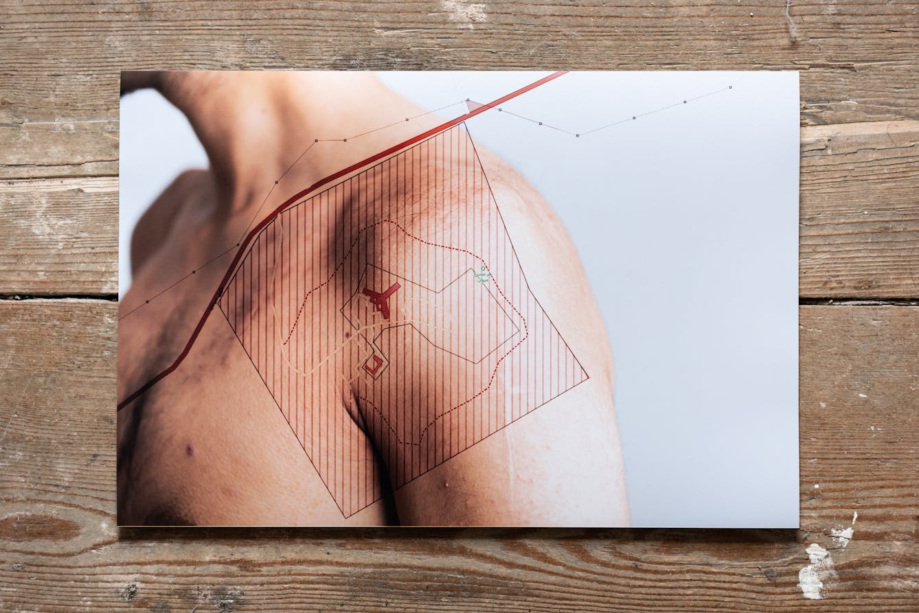 Photograph of a photographic print on a weathered, paint spattered wooden floor. The print shows a naked human shoulders against a white background. Superimposed on shoulders is a simple line map in red, showing the outline of roads and buildings.
