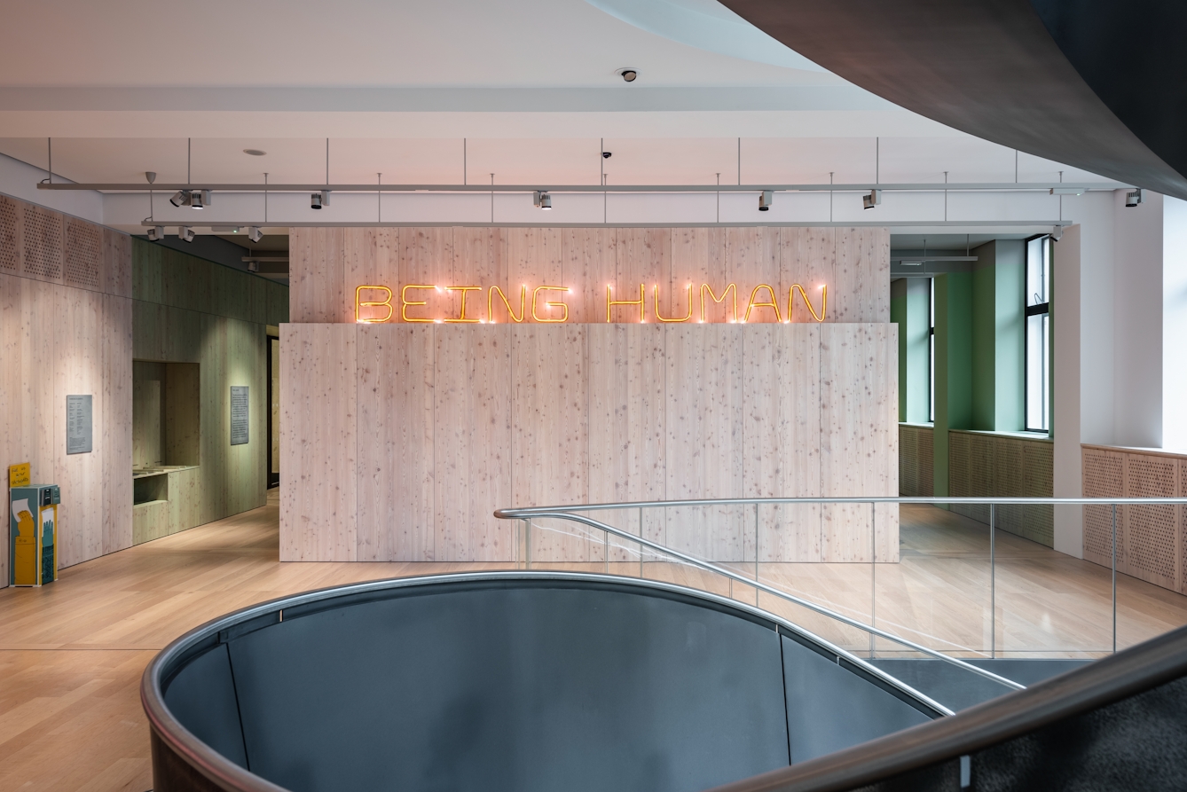 A photograph of the entrance to Being Human as seen from the first floor. Open entrances without doors to the exhibition are on both sides of a central wood panelled wall with a neon orange sign hanging horizontally on it.