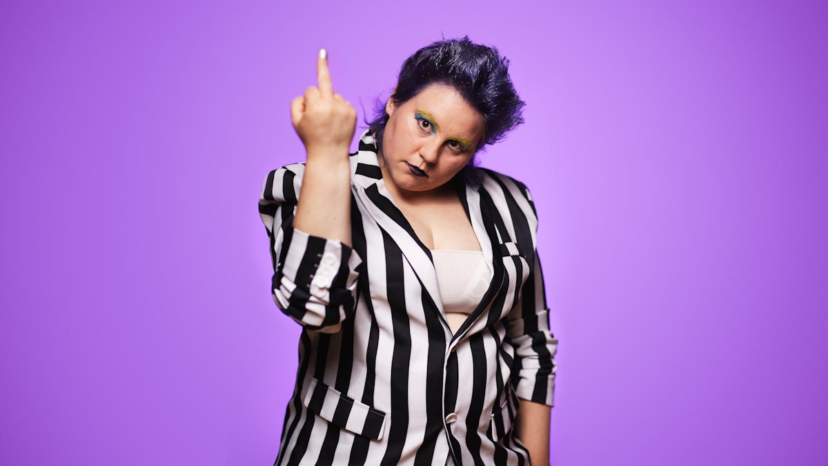 Photograph of a performer dressed in a black and white vertical striped suit. Their sleeves are pulled up to show their forearms. Their right arm is raised up in front of them showing the middle finger of their hand to the camera in an act of defiance. Their hair has a blue tint and their eyes are made up with blue and yellow make-up. They are looking hard, straight into the camera. Behind them, the background is a graduated purple tone.