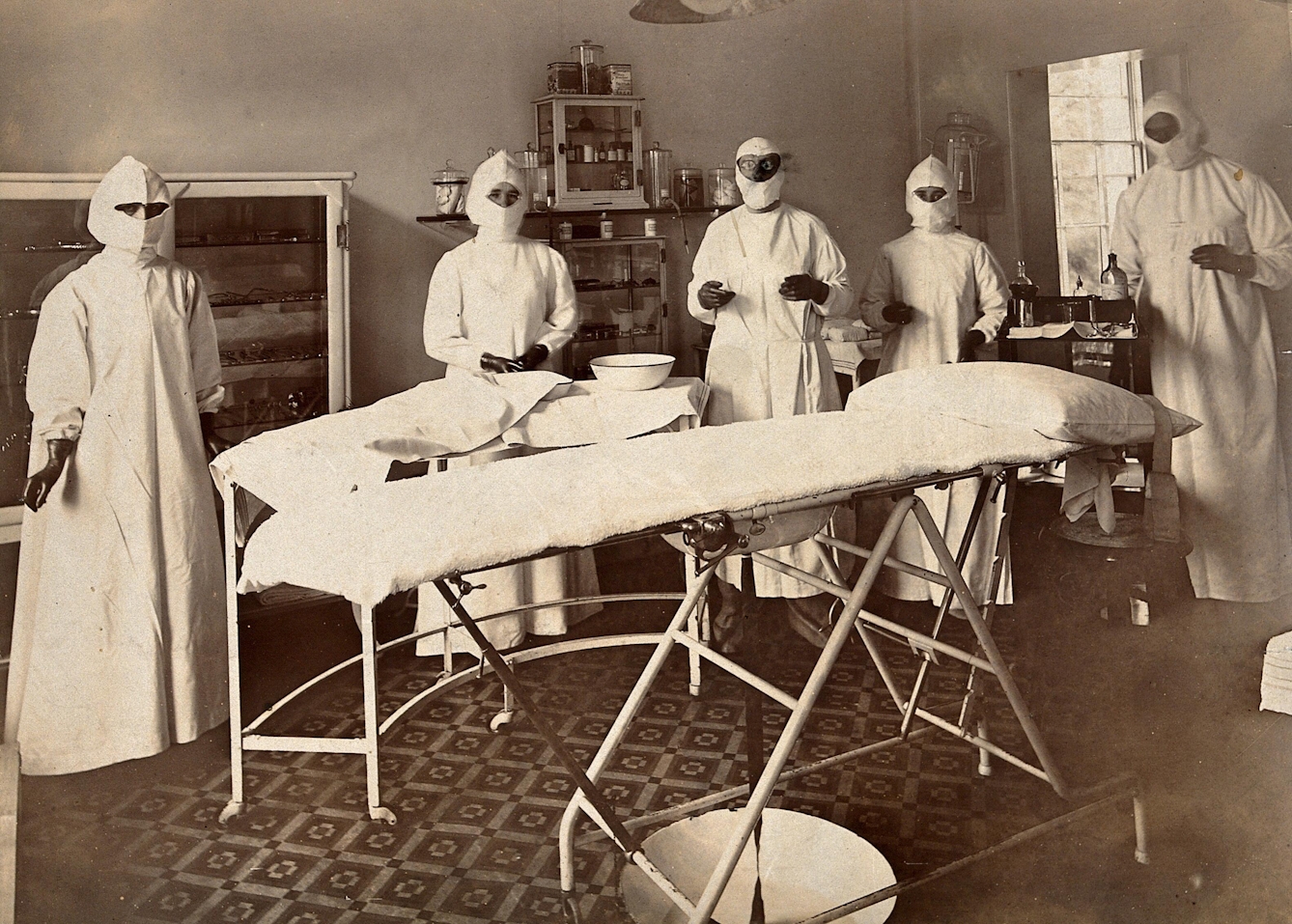 Sepia photograph of an operating theatre in a small tool with various cabinets and apparatus in the background and five medical professionals wearing robes, gloves, and with their faces and hair covered.