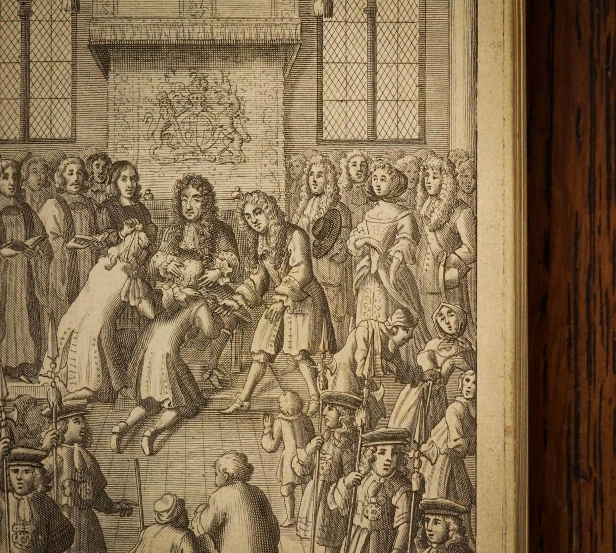 A photograph of an open book against a dark wood background. The page of the book shows an illustration depicting a crowded official ceremony with the King presented with diseased men who are kneeling in front of him.
