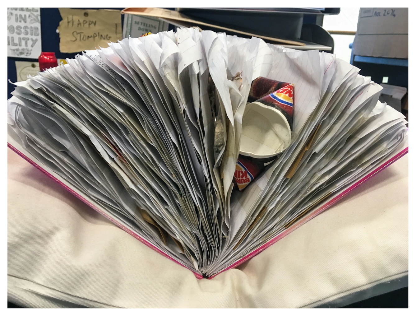 Photograph showing a spiral bound scrapbook resting on a white cushion. The scrapbook is filled with material such that the pages are fanning out under the pressure A squashed paper Burger King drinks cup can be seen crush within the pages. In the background are glimpses of an office space, with boxes and paper sheets pinned to a board.