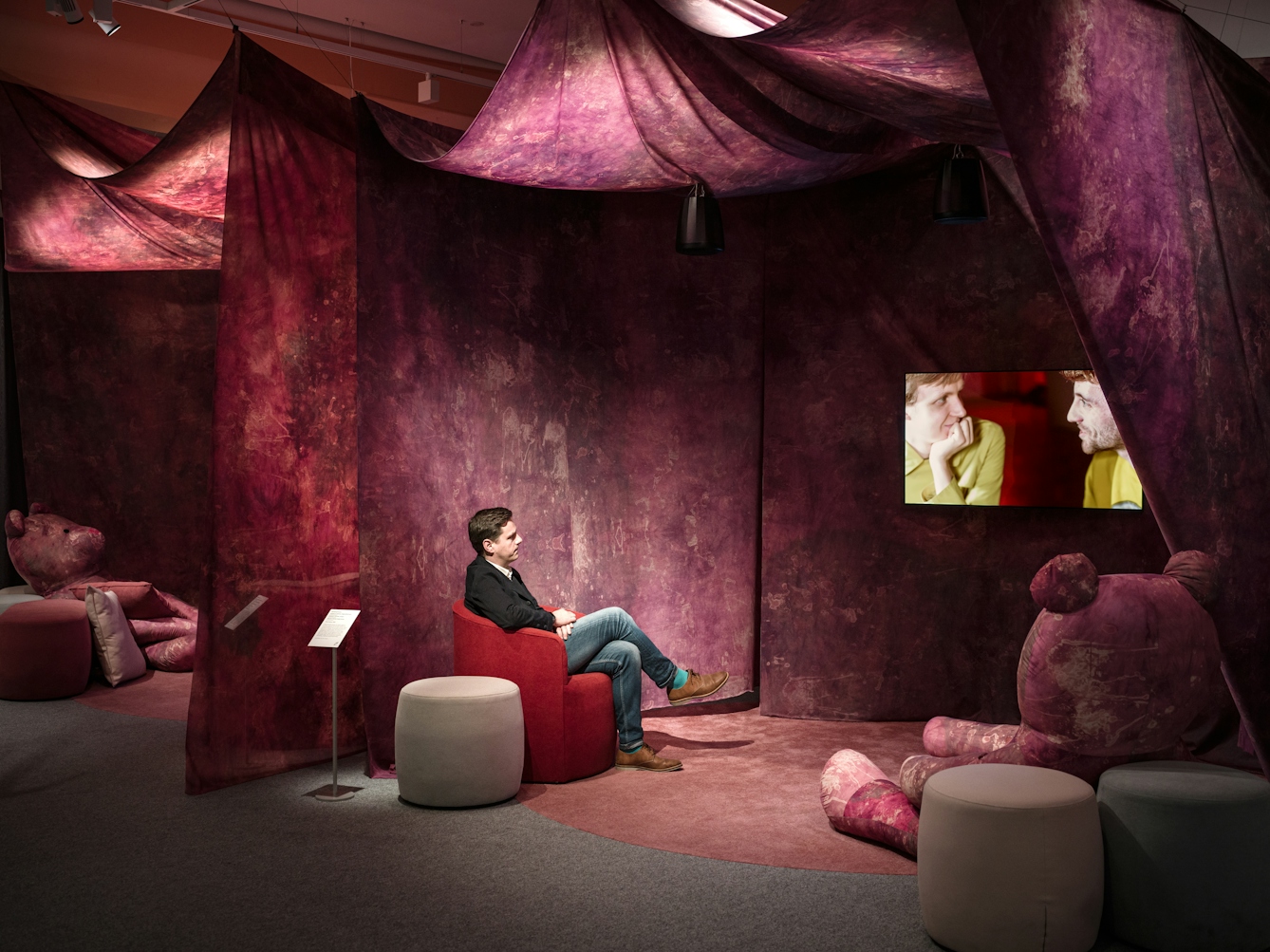 Photograph of a man sitting in a red chair under a purple and red tie-dye 'tent' installation suspended from the ceiling.  The man is looking at a flat panel TV that is hanging from the wall..  Inside the tent are several textile stools and two life size purple and red tie-dye bears.