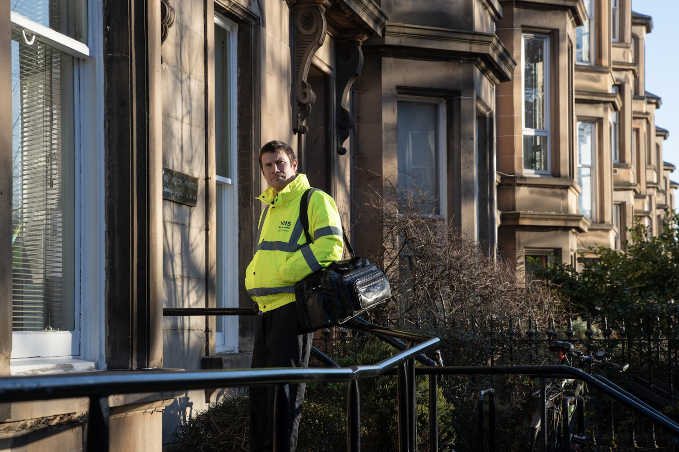 Photograph of a man wearing a hi-viz jacket with 'NHS Scotland' written on it and black trousers. He is walking into the front door of a sandstone building. Over his left shoulder is a large black bag. In the foreground are the black handrails of the entrance steps. Int he background the other buildings on the street can be seen receding into the distance. He is looking to camera as he walks. In front of him is a brass plaque on the wall and some of the words can be seen, 'Medical Practice'.