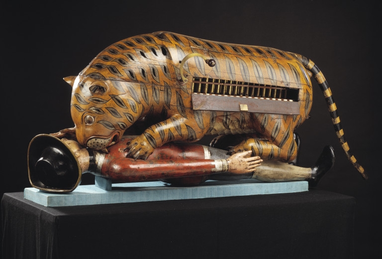 Photograph of Tipu's Tiger, a decorative wooden object. The painted wooden object depicts a European man being savaged by a tiger. The man whose origins are confirmed by his clothes and hat lies on his back while the tiger sinks its teeth into his neck. There is a panel in the tiger's back through which organ pipes are visible. There is a wooden handle next to this. 