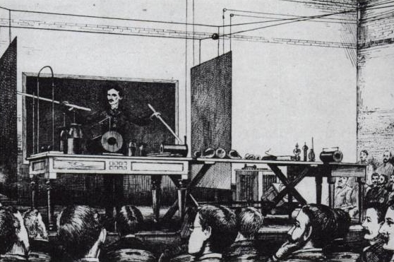 Illustration of Nikola Tesla at the front of a room demonstrating to crowds by pointing at equipment using sticks. 