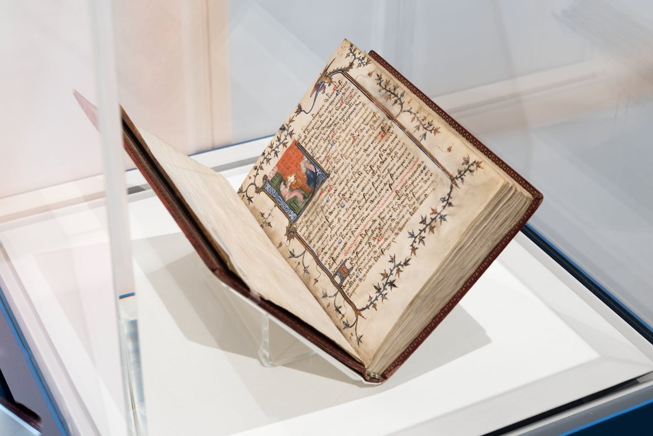Photograph of an exhibition glass display case showing an open manuscript, held in a book cradle. The open double page spread shows intricate writing and page pictorial embellishment on the right hand page with a colourful illumination at the start of the text.
