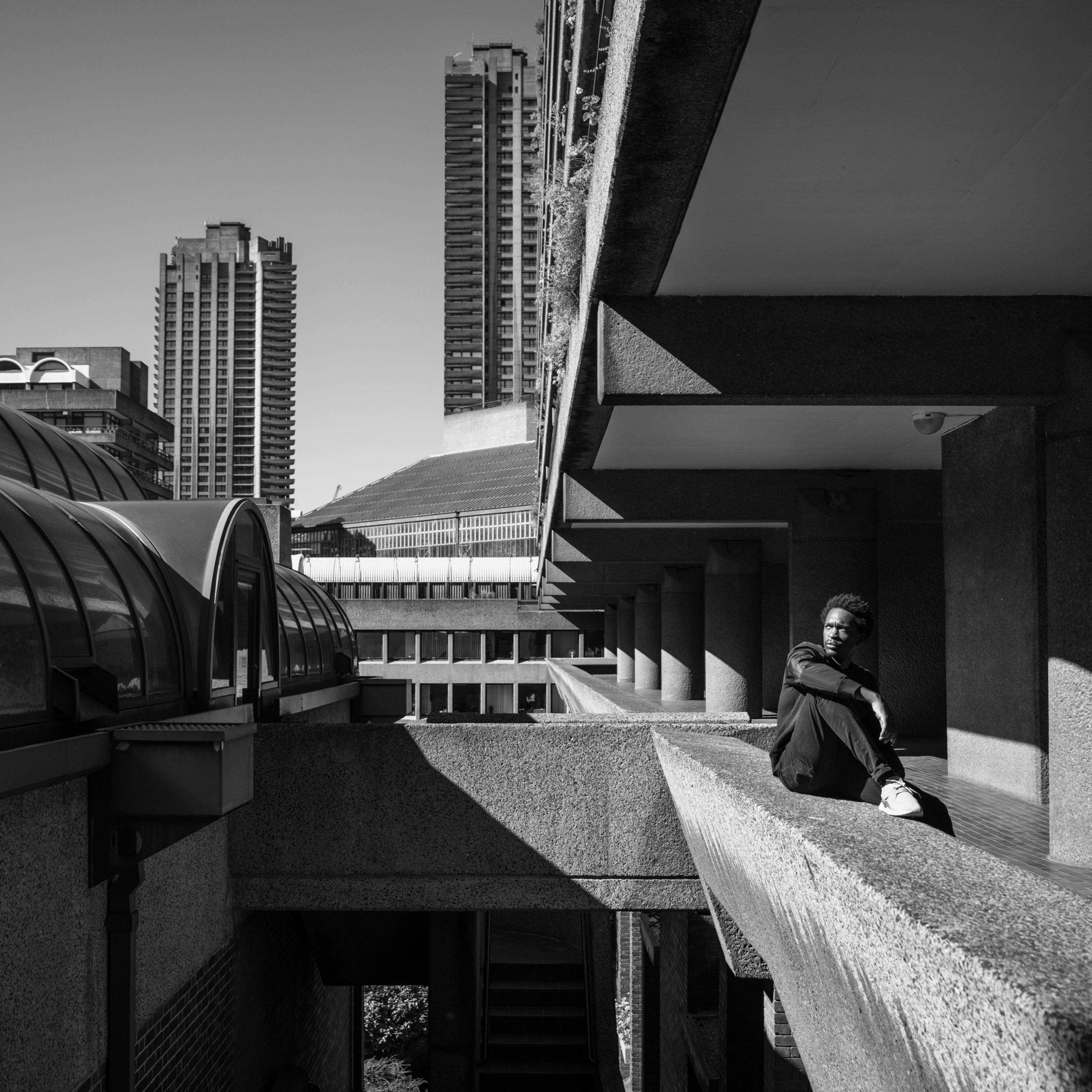 Photograph in black and white of a man sitting on a walkway balcony looking out at the Barbican Centre, London.
