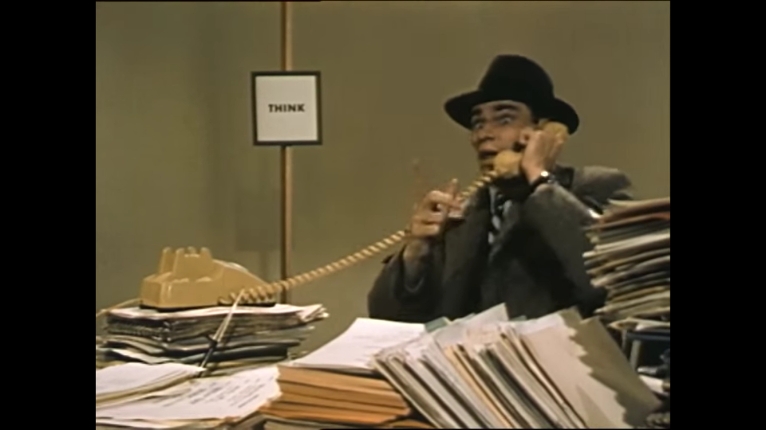 Still image from film of a man wearing a trilby hat talking on the phone with wide-eyes. He is surrounded by toppling piles of paperwork.