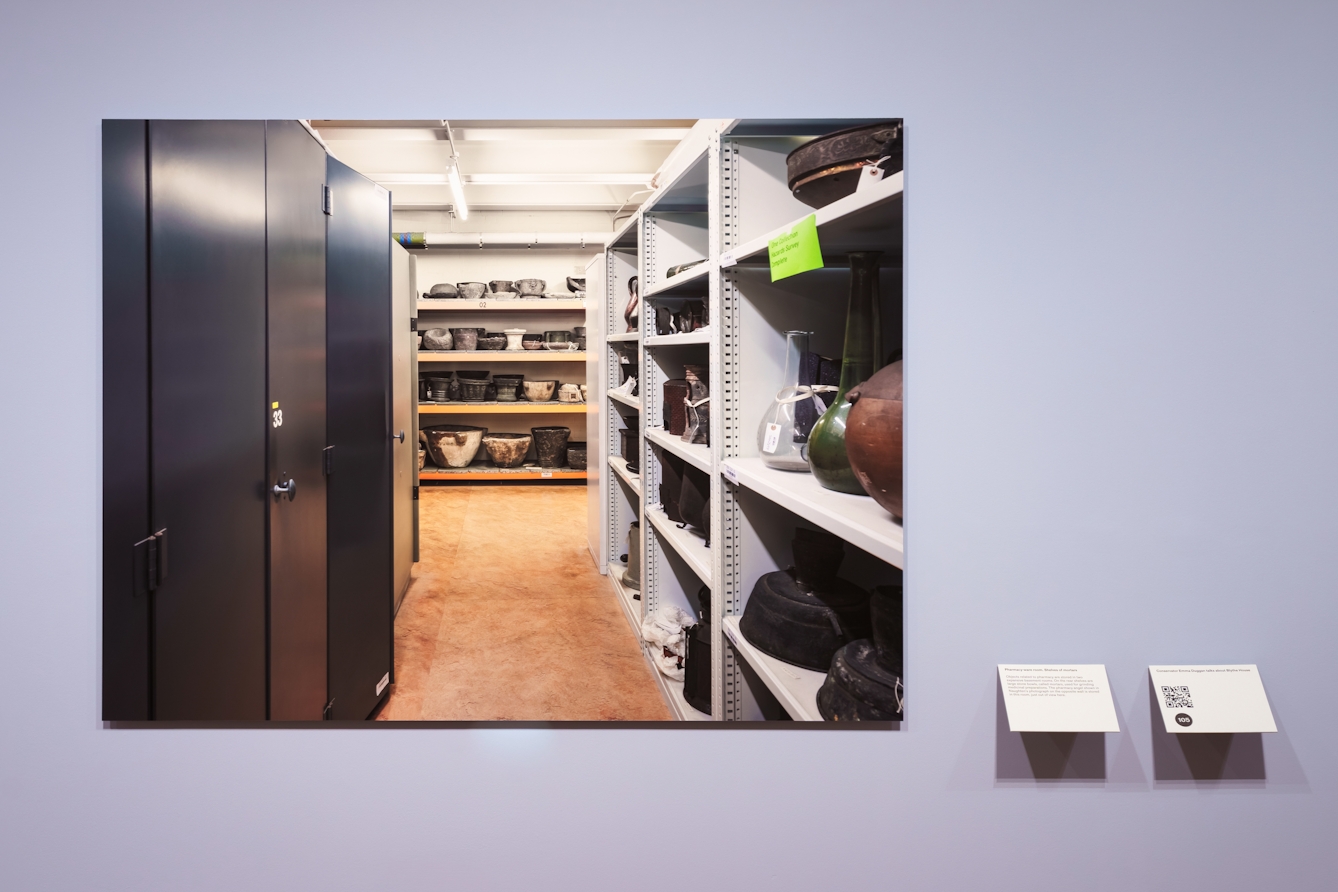 Photograph of a gallery exhibition space showing a large photographic print mounted on the wall with smaller wall mounted information panels. The colour of the wall is light purple. The print shows a museum storage space complete with cabinets, shelving and objects.