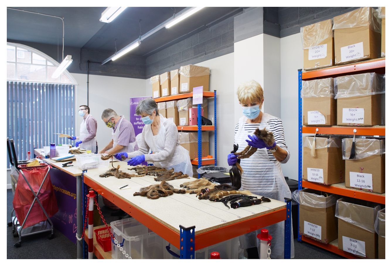 Photograph showing an indoor setting with a large industrial table and shelving coloured blue and orange. Behind the table are 4 individuals wearing white plastic aprons, face coverings and purple latex groves. They are each opening and sorting packages containing cut hair donations. On the tabletop are many groups of cut hair, ties or plaited together. There is a variety of hair coloured and types. On the shelves behind the people are large labelled cardboard boxes.