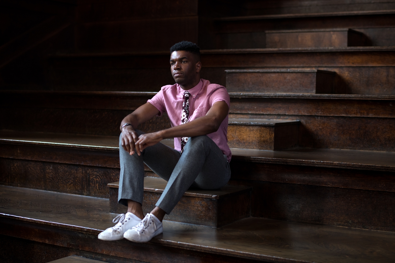 Photograph of a man sitting on the steps of an old, dark wood lecture theatre, wearing a pink shirt, floral tie, grey trousers and white plimsoles. He is looking off camera, into the distance.