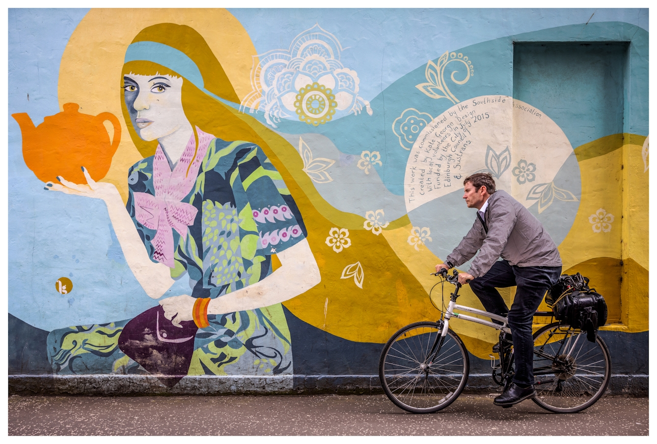Photograph of a doctor on a bicycle cycling past a large painted colourful mural showing a woman with flowing hair holding an orange teapot. He has is large medical bag strapped to his bike rack.