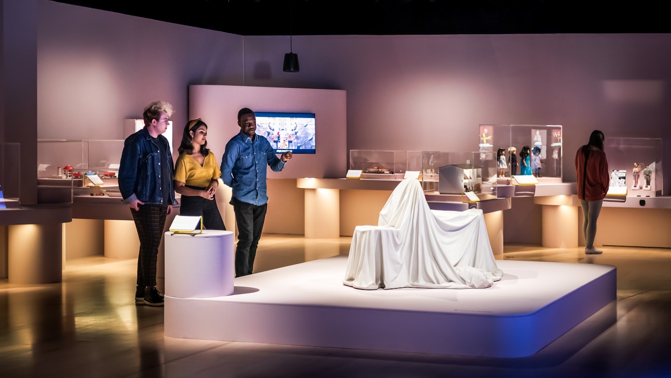 Photograph of visitors exploring the exhibition, Play Well at Wellcome Collection. The photograph shows three people standing next to a low display plinth discussing the exhibit which consists of pieces of furniture covered by a white sheet. Behind them are other display cases against a pink wall and another gallery visitor.