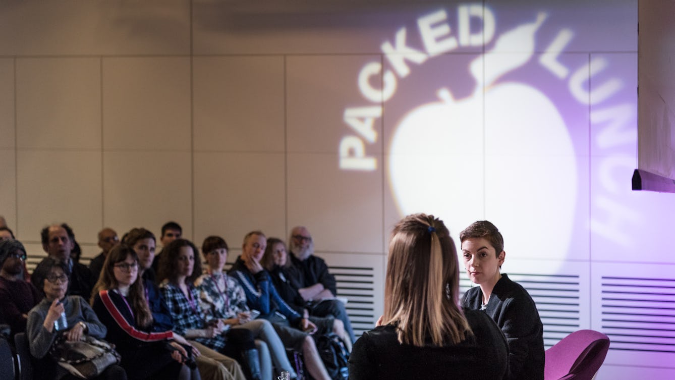 Photograph of two women having a conversation on a small stage. In the background are the audience. Projected onto the far wall is the silhouette of an apple with the words Packed Lunch circling the top of the apple.