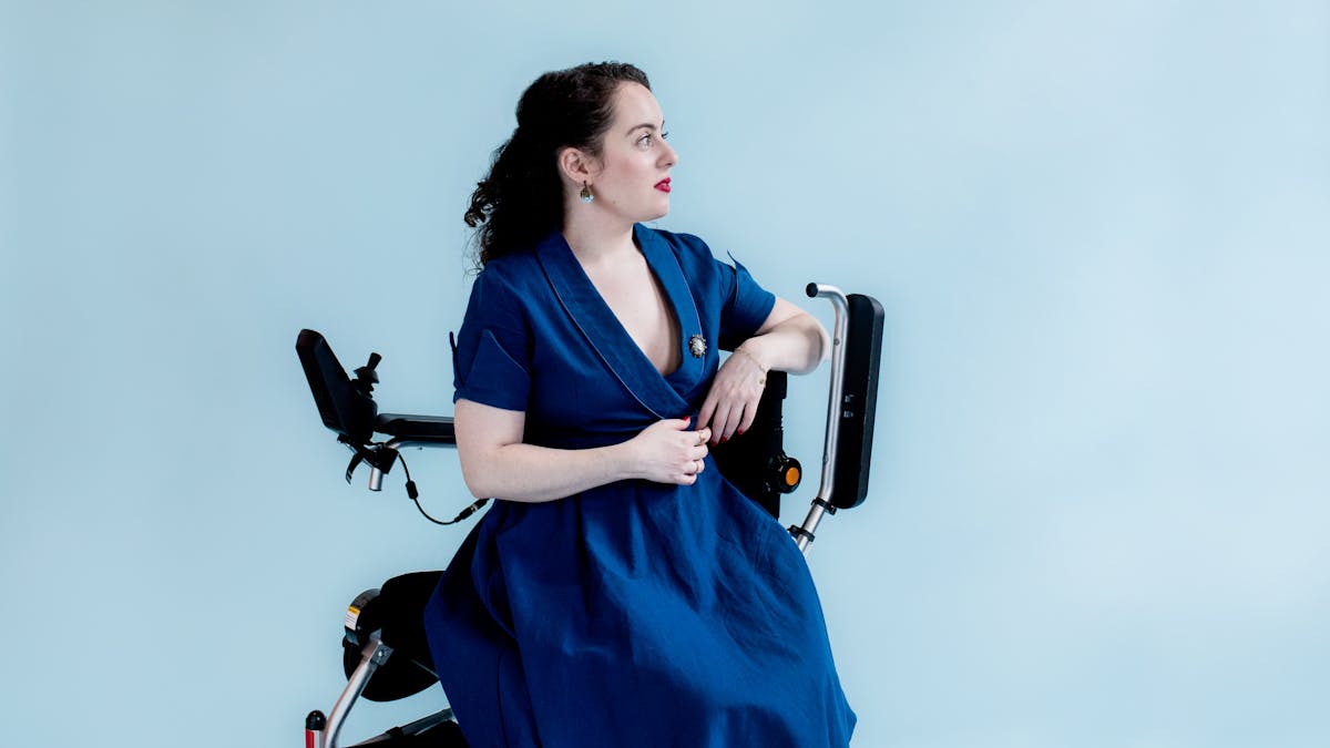 Photograph of a young woman in a blue dress with a broach sitting side-on in her wheelchair against a light blue background. She is looking off to the right of camera, face in profile.