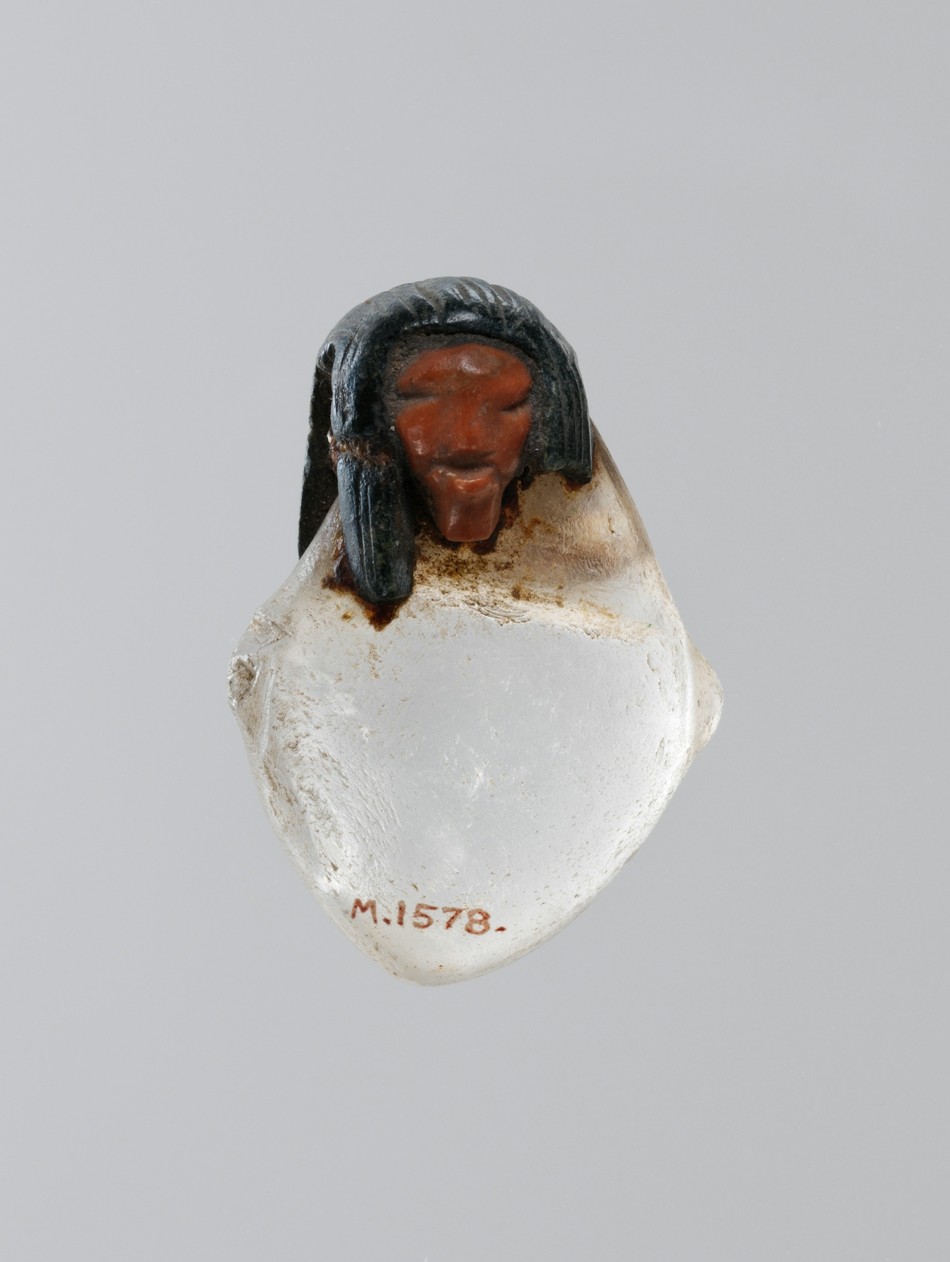 Photograph of a heart shaped amulet with a carved human head, made out of rock crystal, set against a grey background.