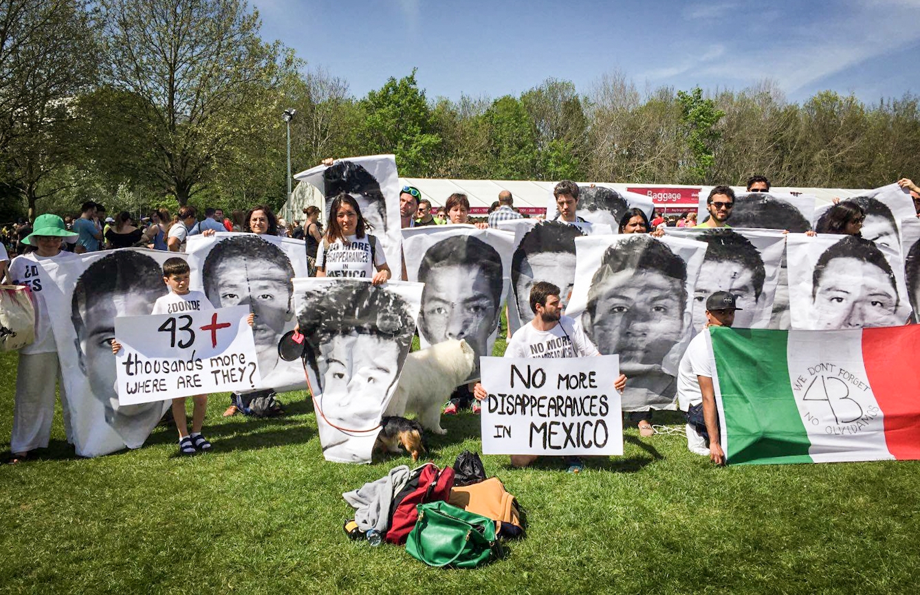 Photograph of a young woman standing in a park, in a sea of protest banners, bearing the faces of people who have disappeared in mexico.