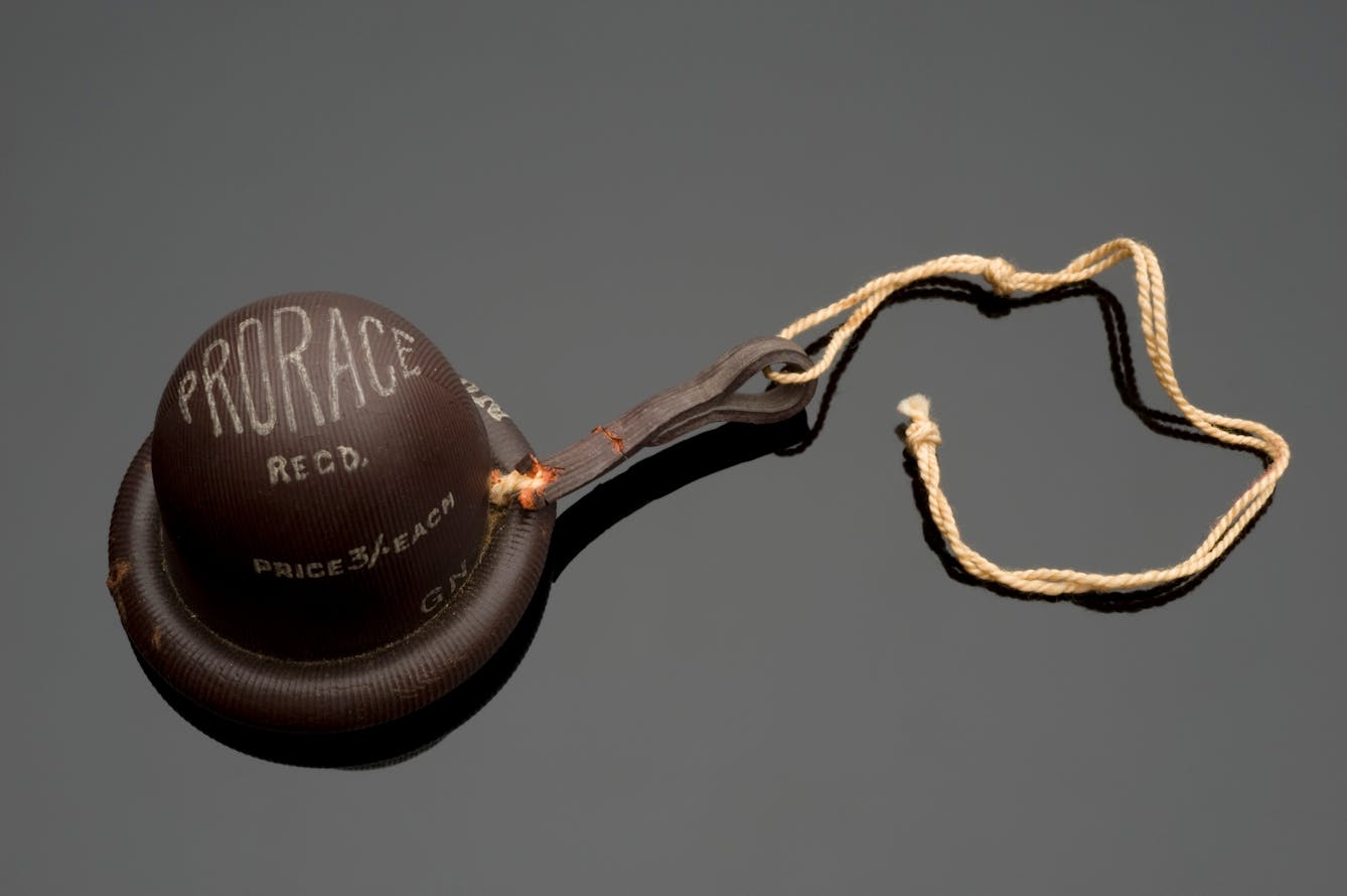 Photograph of a 'Prorace' cervical cap, England, 1915-1925. This brand of cervical cap was a modified version of a French design. Dr Marie Stopes (1880-1958) adapted the design to incorporate a higher dome. She believed this encouraged the cap to maintain its shape with the body. Brown cap with "Prorace" brand imprinted on top, twine handle, on a grey background.