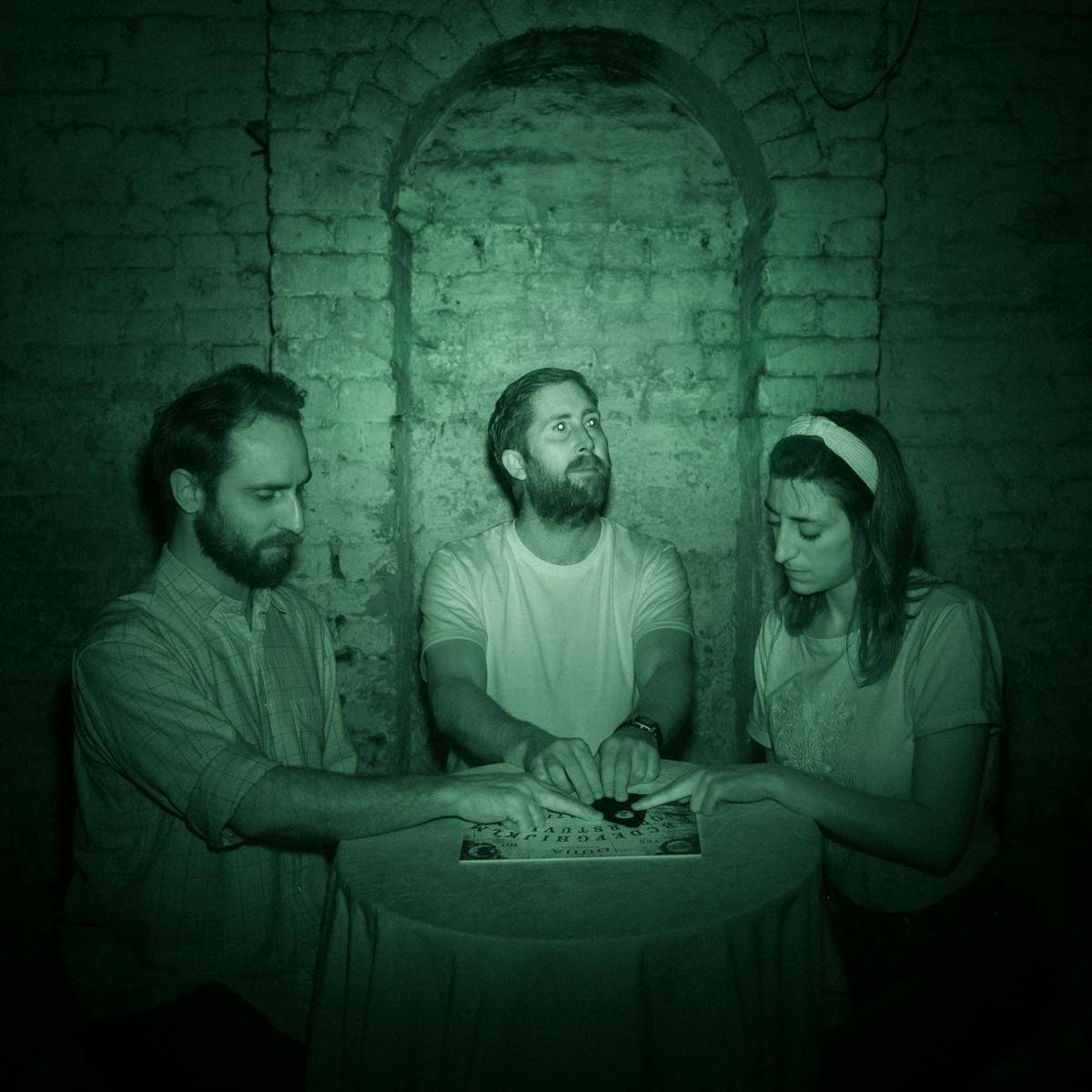 Photograph of three people sat at a round table in a cellar. Behind them is a white brick wall with an arch. Their hands rest on the planchette of a ouija board. The image is toned green as a result of being made under infrared light.